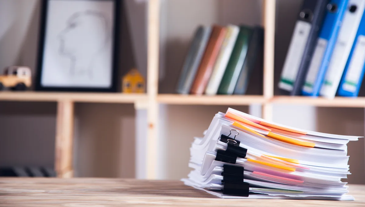 A backlog of paper files stacked on an office desk